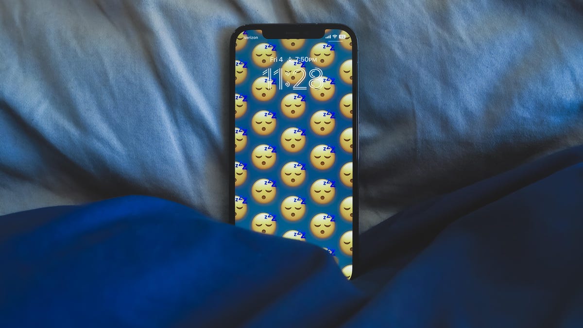 An iPhone sits tucked into bed beneath pillow and comforter.