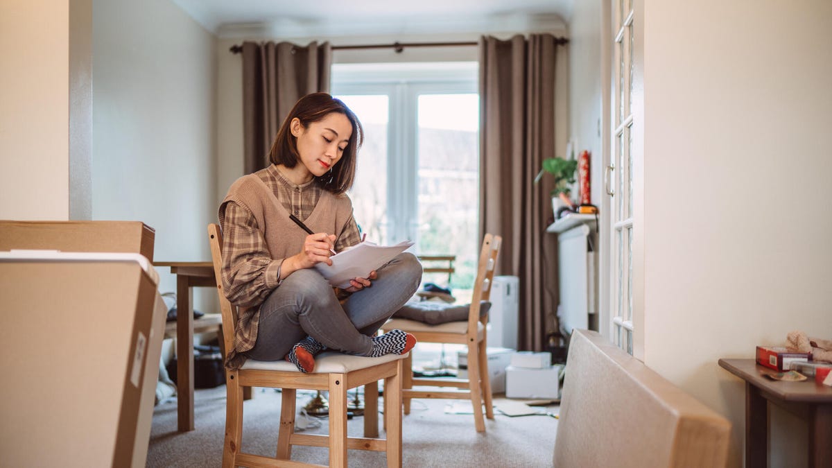 A woman makes notes on a chair in a house surrounded by moving boxes.