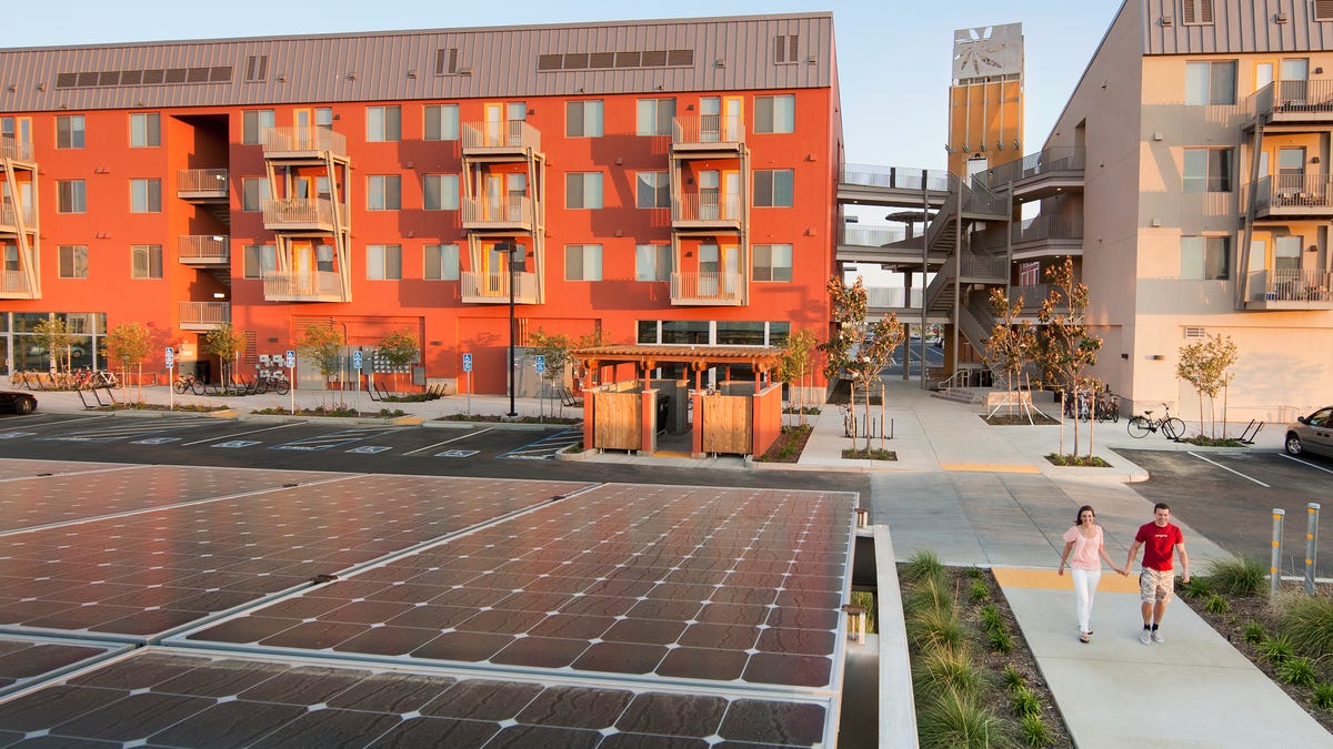 Two people walk by a large solar array in front of an orange apartment building