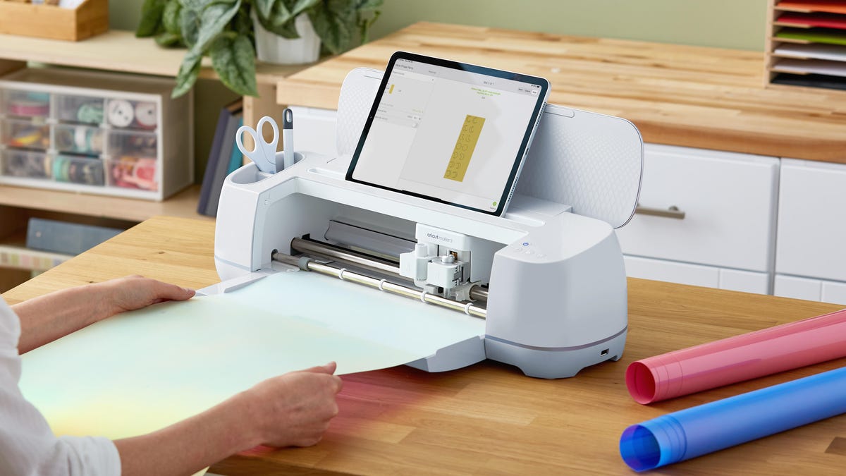 A white Cricut Maker 3 sitting on a tabletop with a tablet attached to it and a white person's hands feeding material into the machine.