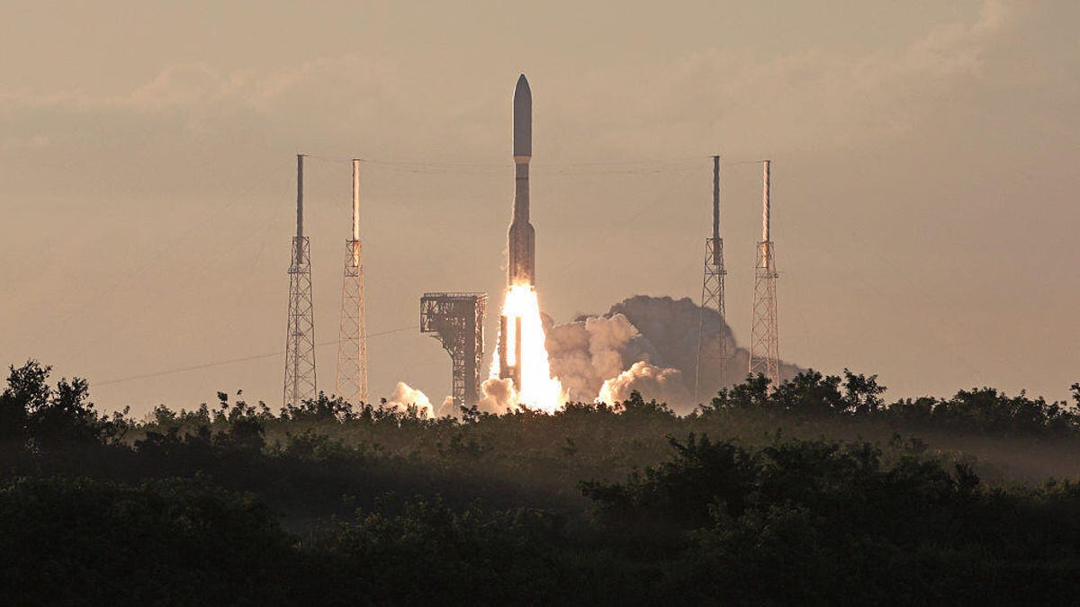 Atlas V rocket launches in the distance, silhoutted trees in foreground