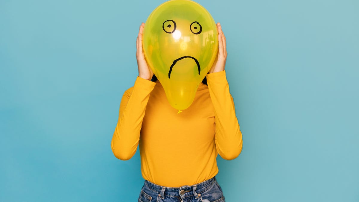 Woman hidden behing a balloon with a sad face drawn on it over blue background.