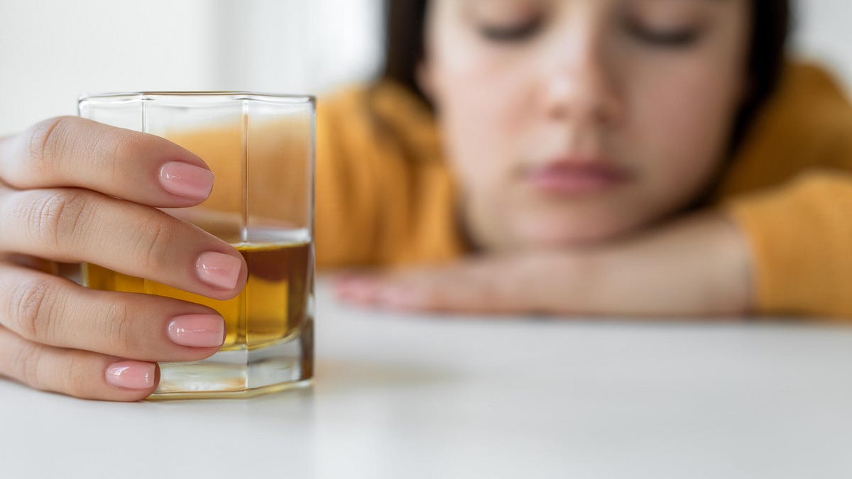A person wearing a yellow shirt with light pink nails leaning on a white surface with a whiskey glass in their hand.