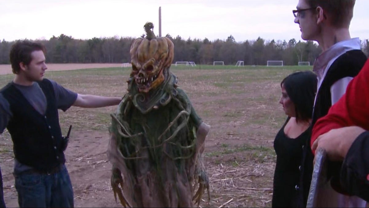 in a still from the movie Hell House LLC, four people stand looking at another person dressed in a scary Halloween costume with an angry pumpkin head