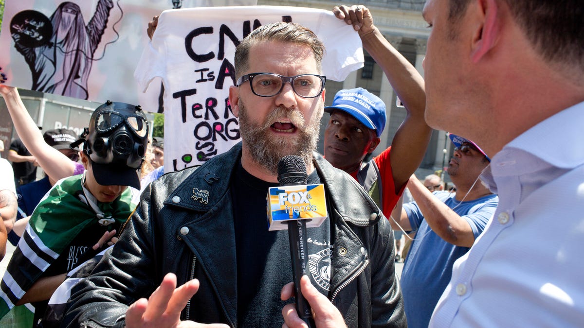 Gavin McInnes answers questions from a journalist in front of rally goers holding signs.