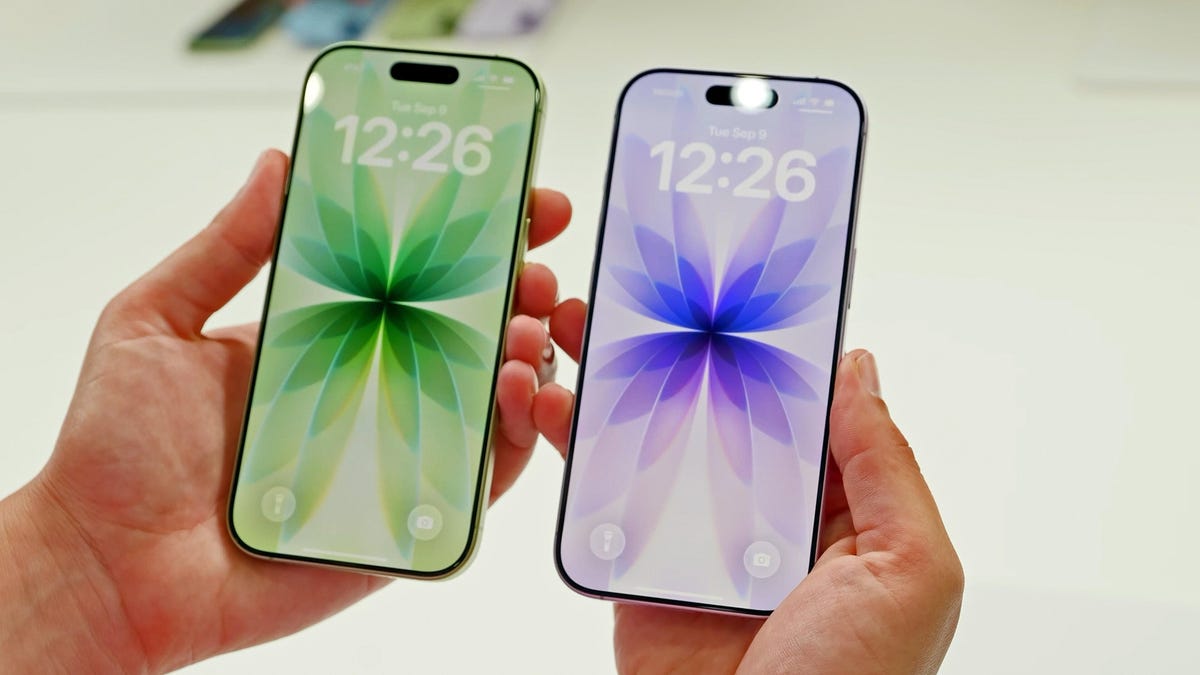 Two iPhone 17 phones, one green and one purple, being held in a pair of hands against a light background.