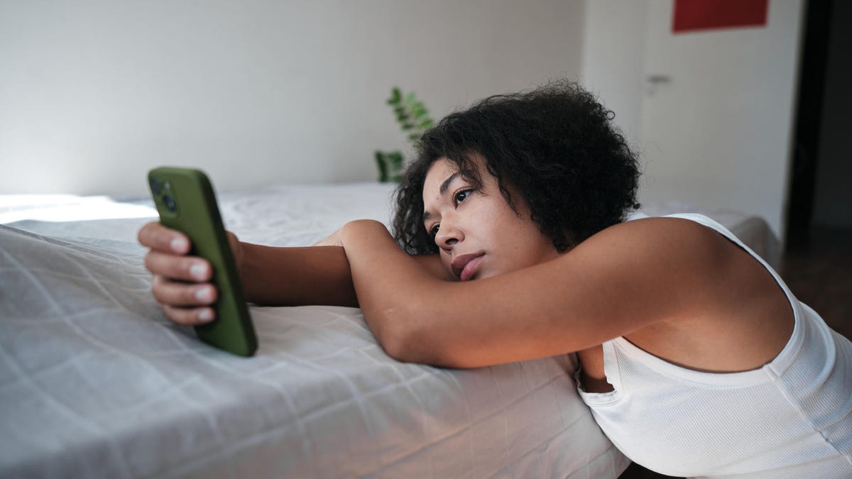 A person with short curly black hair leaning on a white bed and looking at their phone with a sad expression.
