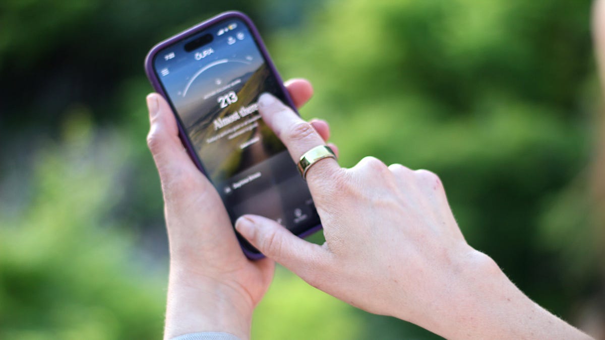 The fingers of the hand have navy nail polish, and the index finger is wearing a navy Oura Ring. The background is light blue.