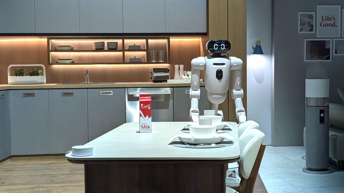 CLOiD stand in a kitchen. The white robot waits next to a table that is set for dinner for the household humans.