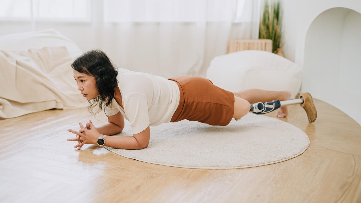A woman with black, shoulder-length hair, a white tee, brown shorts, and a prosthetic leg doing a plank on the floor.