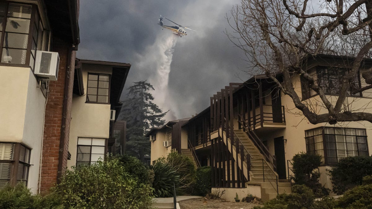 Framed by an apartment building and its entry court in the foreground, a swooping helicopter dumps water, with the falling liquid set off against a sky filled with smoke.