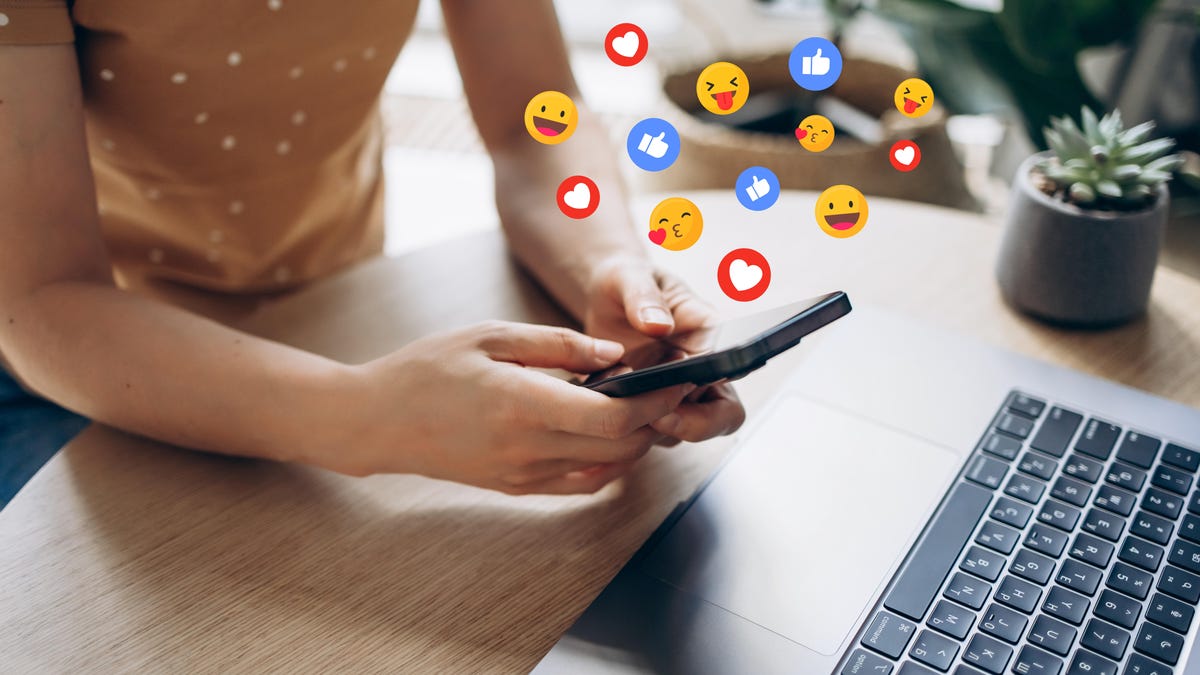 Woman holding a cellphone in front of a laptop with emojis floating in the air