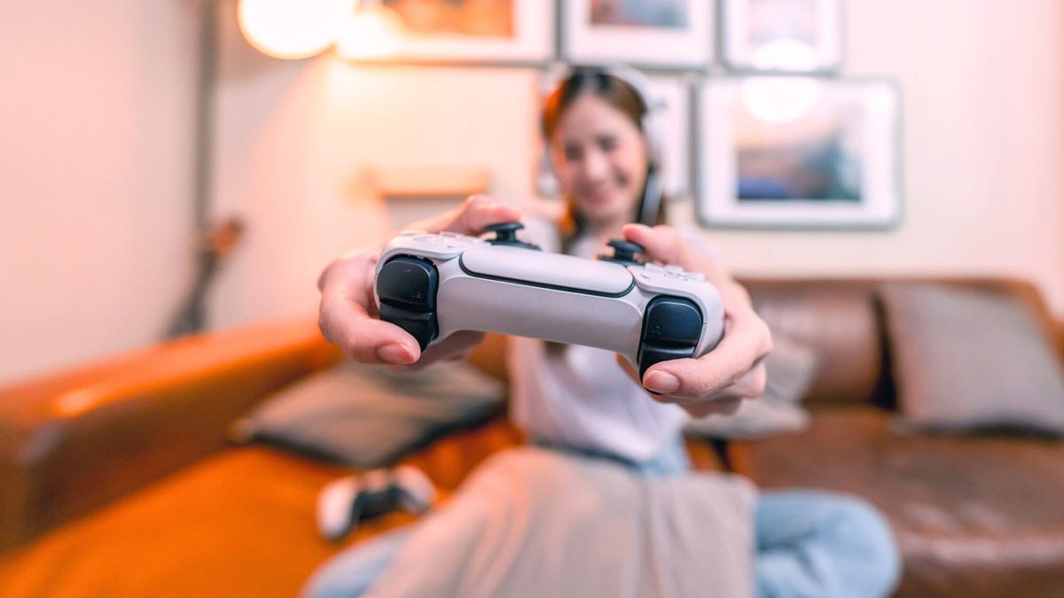 A photograph shows a smiling woman holding a gaming console out toward the camera, her thumbs on the buttons. The console is in the foreground, in sharp focus, and the woman is blurred.