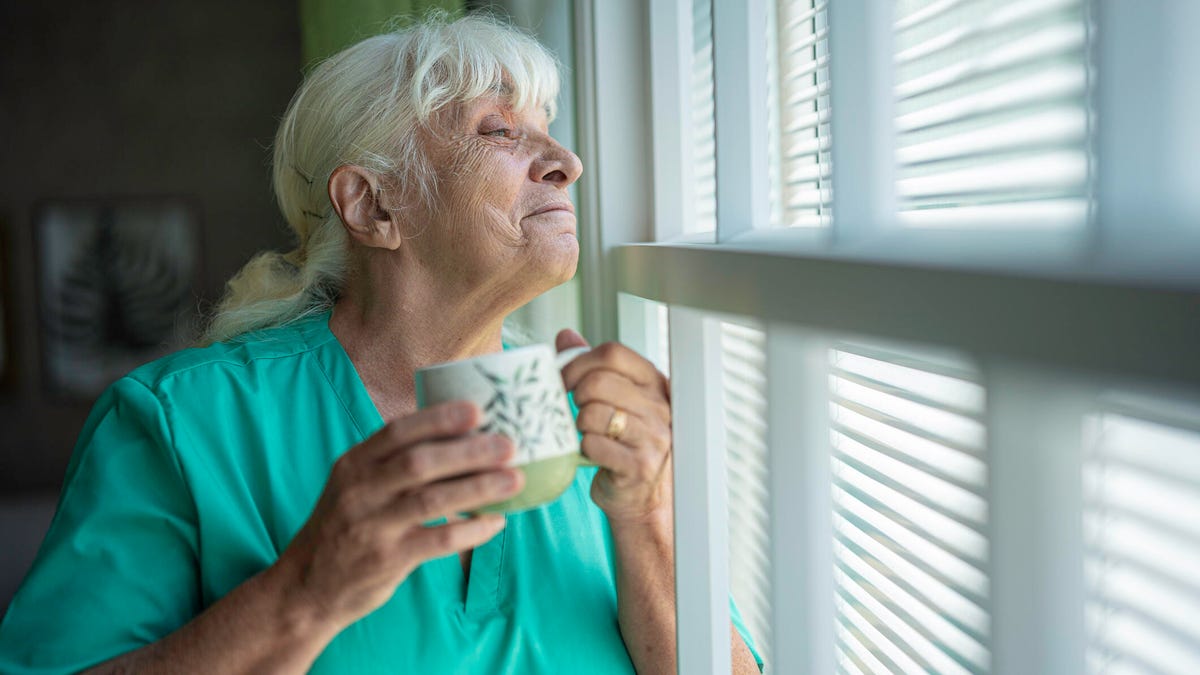 An old woman looks happily out through window blinds while holding a cup.