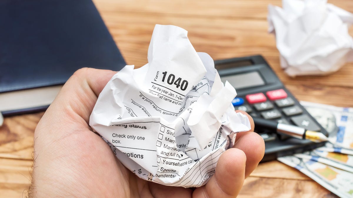 A hand holds a crumpled Form 1040 tax form over a light brown wooden desk holding a calculator, hardback book, and several hundred-dollar bills